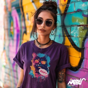 Woman wearing sunglasses and a purple t-shirt. The t-shirt has a graffiti style cat who's also wearing sunglasses. There is a speech bubble coming from the cat with the text 'Sup?'