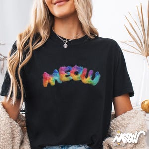 K&ouml;nnte beinhalten: Schwarzes T-Shirt mit dem Wort "MEOW" in einem bunten, regenbogenartigen, flauschigen Schriftzug. Das Shirt hat einen Rundhalsausschnitt. Das Bild zeigt auch eine Frau, die eine Halskette tr&auml;gt.