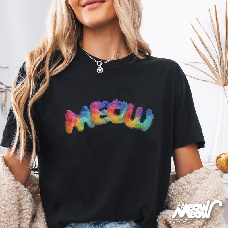 K&ouml;nnte beinhalten: Schwarzes T-Shirt mit dem Wort "MEOW" in einem bunten, regenbogenartigen, flauschigen Schriftzug. Das Shirt hat einen Rundhalsausschnitt. Das Bild zeigt auch eine Frau, die eine Halskette tr&auml;gt.