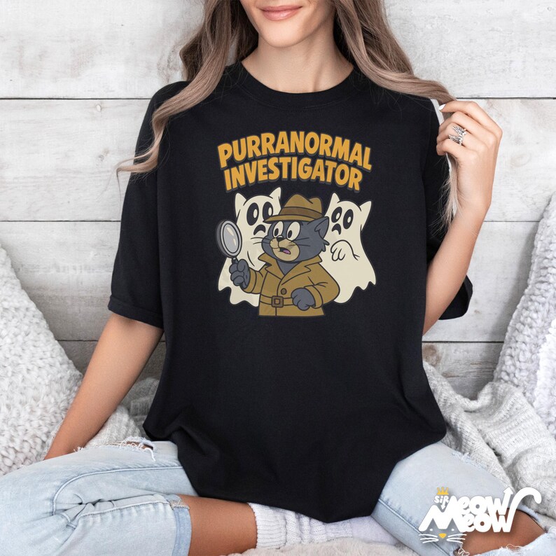 May include: Black t-shirt featuring a cartoon cat detective with a magnifying glass, wearing a trench coat and hat, with two ghost figures. The text "PURRANORMAL INVESTIGATOR" is above the cat.