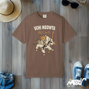 K&ouml;nnte beinhalten: Ein braunes T-Shirt mit einer Grafik von zwei Katzen in wei&szlig;en Kampfkunst-Uniformen mit schwarzen G&uuml;rteln, die einen Judo-Wurf ausf&uuml;hren. Der Text UCHI MEOWTA und japanische Schriftzeichen sind in hellem Creme &uuml;ber den Katzen gedruckt.