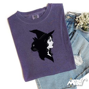May include: A purple Comfort Colors t-shirt featuring a black graphic of a witch's silhouette combined with a black cat. The witch is wearing a pointed hat. The shirt is folded on top of a pair of denim shorts.