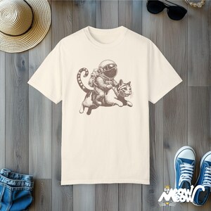 Ivory Comfort Colors 1717 unisex T-shirt with a humorous white graphic of an astronaut riding atop a playful cat, combining outer space and cat lover themes.