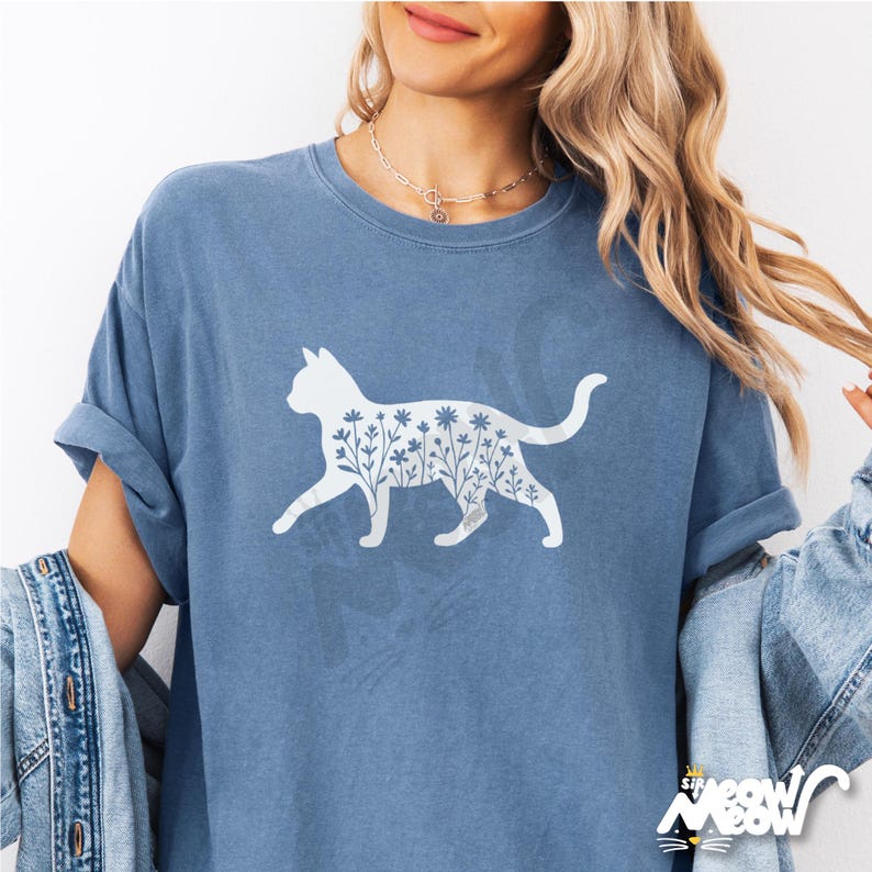 May include: A blue t-shirt featuring a white cat silhouette filled with floral designs. The shirt has rolled-up sleeves and a relaxed fit. The text "sip meow" is at the bottom right corner.