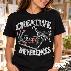 May include: A black short-sleeve t-shirt featuring a graphic of a tabby cat walking across a canvas. The text reads CREATIVE DIFFERENCES. The design includes three paintbrushes, a mountain landscape, and red paw prints on the canvas.