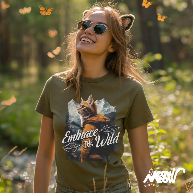 May include: A woman wearing a green t-shirt with a graphic of a cat wearing a backpack and the text "Embrace the Wild".