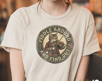May include: Beige t-shirt featuring a circular graphic of a cat playing a violin, dressed in a suit. The graphic reads "Fiddle Around and Find Out" in a vintage style. The shirt has short sleeves.