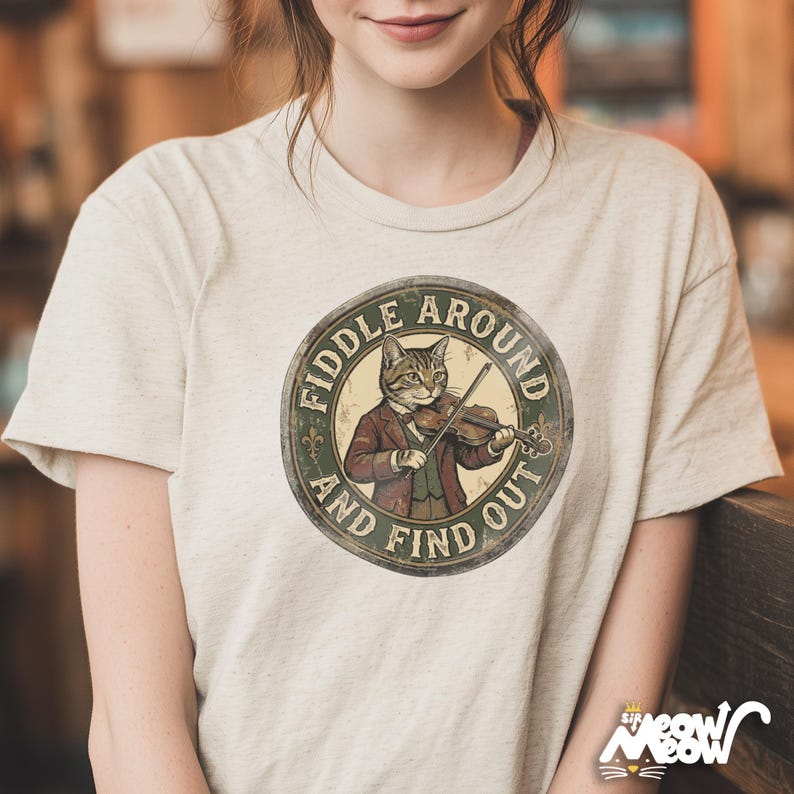 May include: Beige t-shirt featuring a circular graphic of a cat playing a violin, dressed in a suit. The graphic reads "Fiddle Around and Find Out" in a vintage style. The shirt has short sleeves.