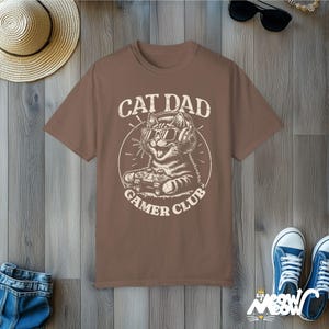 Cat Parent Gamer Club T-Shirts. The tee shows a cat holding a gaming controller. Above the image is the text "Cat Dad" and below the image is the text "Gamer Club".
The t-shirt shown is in the color "Espresso" and is also available in a "Cat Mom" version.
