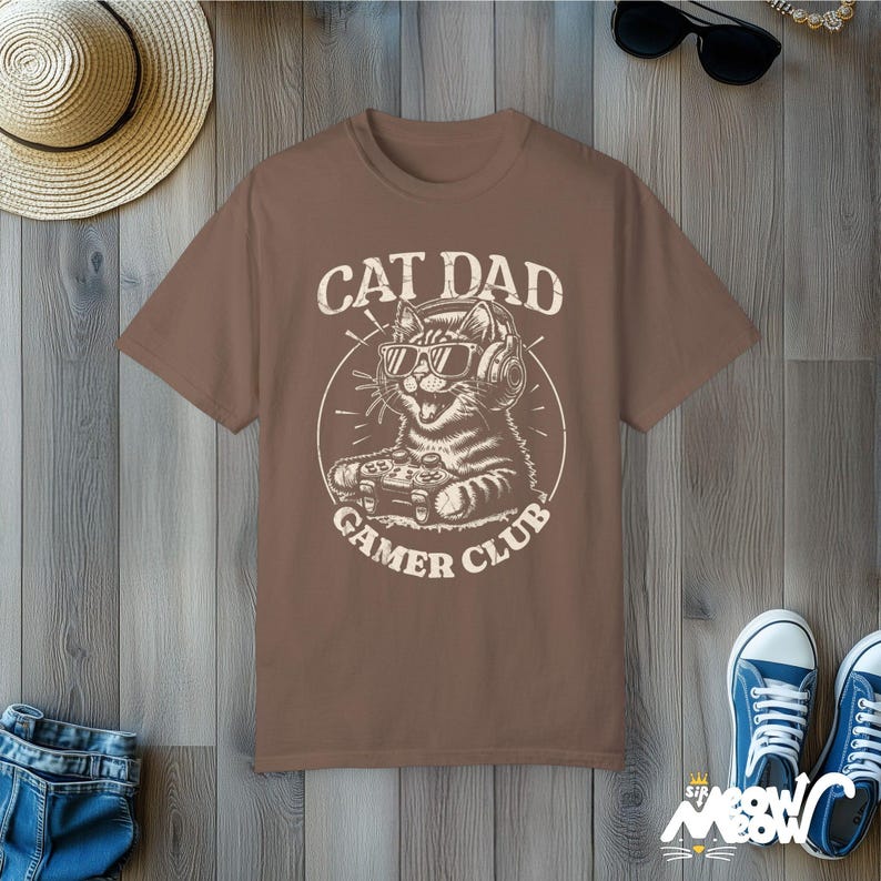 Cat Parent Gamer Club T-Shirts. The tee shows a cat holding a gaming controller. Above the image is the text "Cat Dad" and below the image is the text "Gamer Club".
The t-shirt shown is in the color "Espresso" and is also available in a "Cat Mom" version.