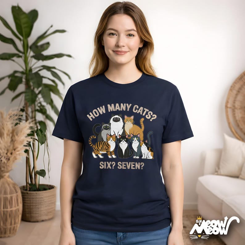 May include: Navy blue t-shirt featuring a graphic of seven cartoon cats with the text "HOW MANY CATS? SIX? SEVEN?" The cats are in various colors and patterns. The shirt is worn by a person.