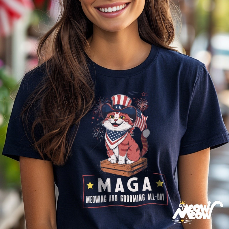 May include: A navy blue t-shirt with a cartoon cat wearing a red, white, and blue hat and bandana. The cat is standing on a wooden crate. The text on the shirt reads "Maga Meowing and Grooming All-Day".