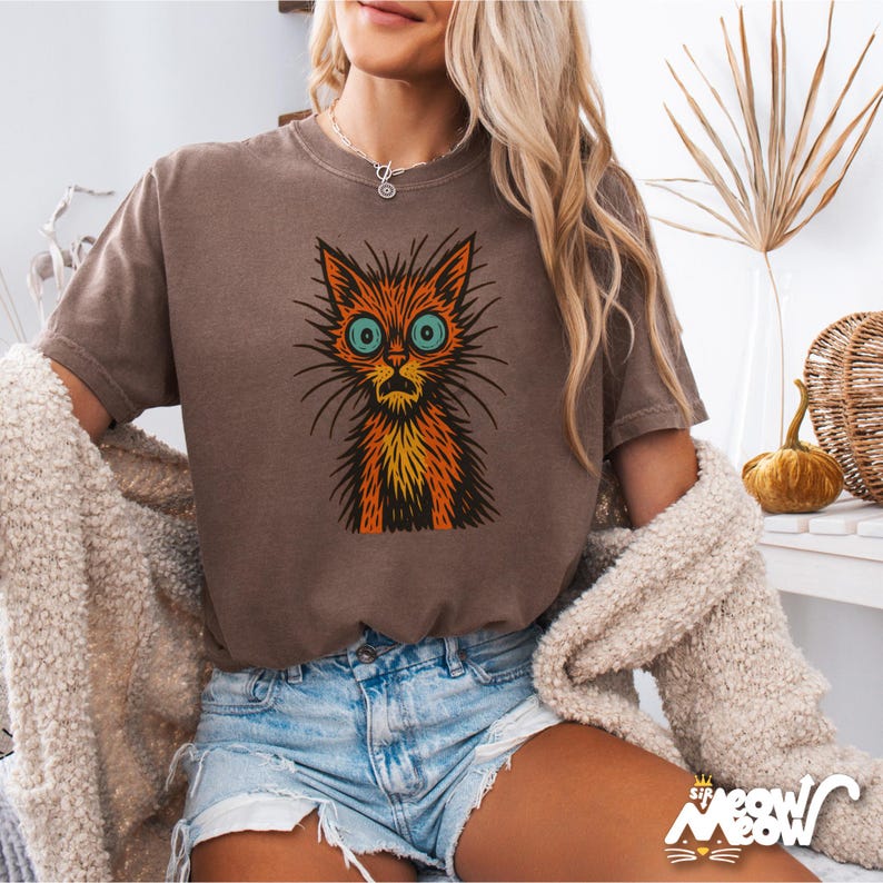 May include: A brown t-shirt featuring a cartoon cat with orange and yellow fur, large blue eyes, and a surprised expression. The cat graphic is centered on the shirt. The shirt is paired with denim shorts and a cream-colored cardigan.
