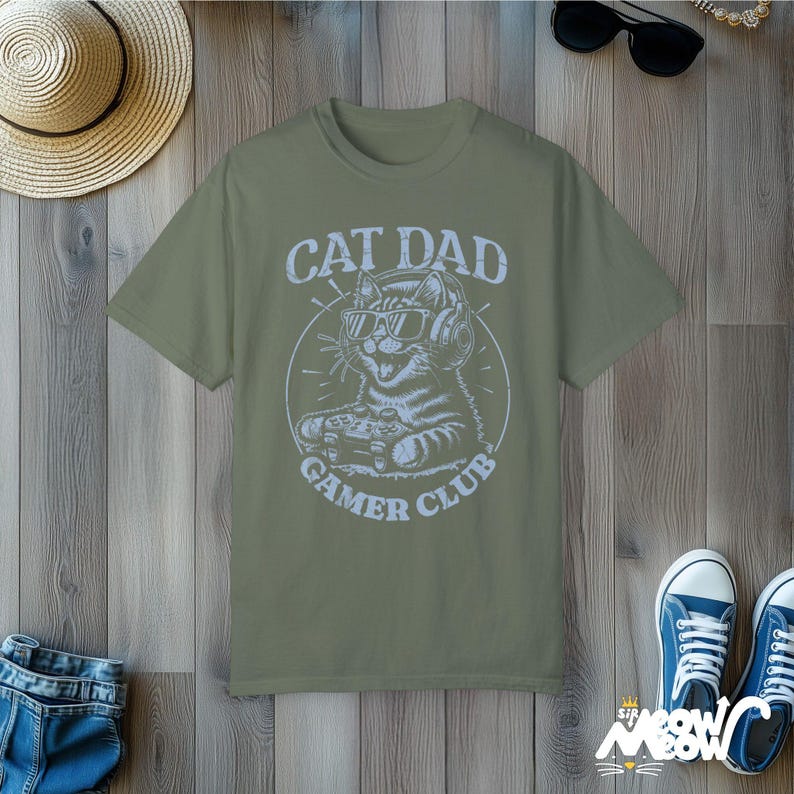 Cat Parent Gamer Club T-Shirts. The tee shows a cat holding a gaming controller. Above the image is the text "Cat Dad" and below the image is the text "Gamer Club".
The t-shirt shown is in the color "Moss" and is also available in a "Cat Mom" version.