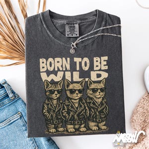 Puede incluir: Camiseta gris oscuro con el texto "BORN TO BE WILD" y tres gatos de dibujos animados con gafas de sol y chaquetas de cuero. Un collar plateado con un colgante est&aacute; sobre la camiseta. La camiseta est&aacute; doblada sobre una superficie blanca.