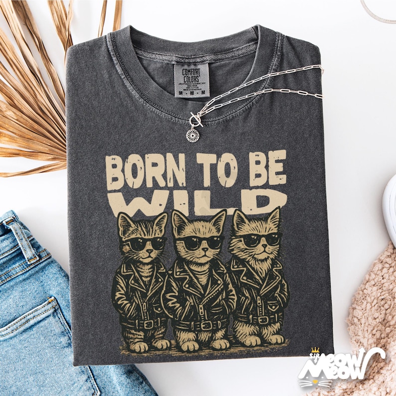 Puede incluir: Camiseta gris oscuro con el texto "BORN TO BE WILD" y tres gatos de dibujos animados con gafas de sol y chaquetas de cuero. Un collar plateado con un colgante est&aacute; sobre la camiseta. La camiseta est&aacute; doblada sobre una superficie blanca.