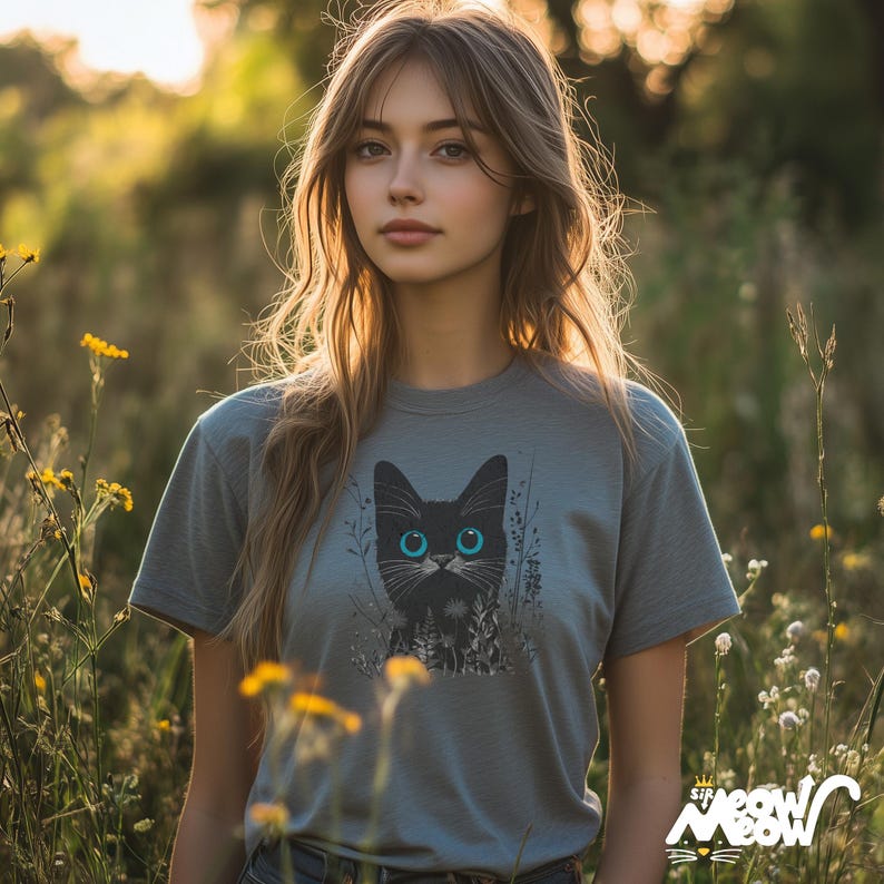 May include: A woman with long brown hair is wearing a gray t-shirt with a black cat graphic. The cat has blue eyes and is surrounded by green foliage. The woman is standing in a field of yellow wildflowers.
