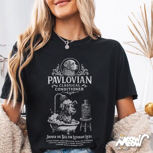 K&ouml;nnte beinhalten: Schwarzes T-Shirt mit einem Grafikdesign mit dem Text "PAVLOVIAN CLASSICAL CONDITIONER" und einer Illustration eines Hundes in einer Badewanne. Das Shirt enth&auml;lt auch den Text "ANSWER THE BELL FOR LUXURIANT LOCKS."