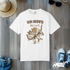 K&ouml;nnte beinhalten: Ein wei&szlig;es Kurzarm-T-Shirt mit einer Grafik von zwei Katzen in Kampfkunst-Uniformen mit schwarzen G&uuml;rteln, die einen Judowurf ausf&uuml;hren. Der Text UCHI MEOWTA und japanische Schriftzeichen sind in Braun &uuml;ber der Katzenillustration gedruckt.