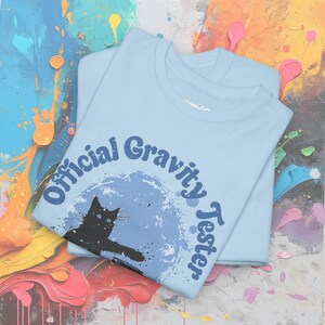 May include: A light blue t-shirt featuring a black cat silhouette and the text "Official Gravity Tester" in a retro font.