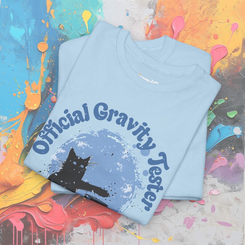 May include: A light blue t-shirt featuring a black cat silhouette and the text "Official Gravity Tester" in a retro font.