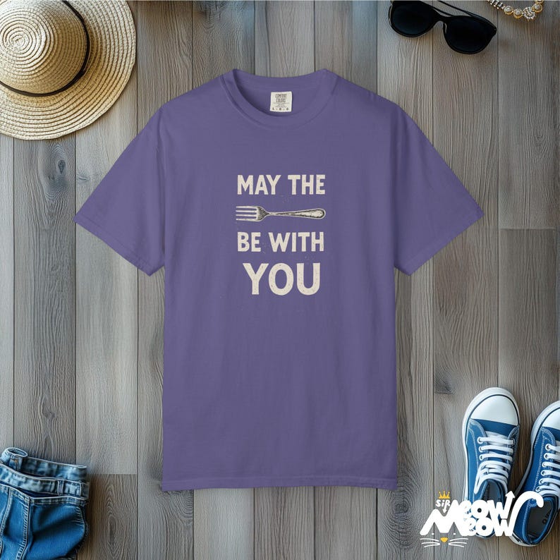 Peut inclure: Un t-shirt violet avec le texte "MAY THE FORK BE WITH YOU" en blanc. Le t-shirt est pos&eacute; sur une surface en bois, avec un chapeau de paille, des lunettes de soleil et des baskets bleues en arri&egrave;re-plan.