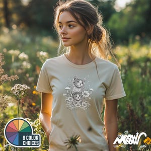 K&ouml;nnte beinhalten: Eine Frau tr&auml;gt ein hellbeiges T-Shirt mit einer Schwarzwei&szlig;illustration von zwei K&auml;tzchen in einer Blumenumgebung. Die K&auml;tzchen schauen den Betrachter an. Das T-Shirt hat den Text "Meow Meow" in der rechten unteren Ecke.