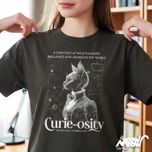May include: Dark gray t-shirt featuring a stylized cat in a suit and bow tie, with the text "Curie-osity" and "A curious cat who radiated brilliance and changed the world." The shirt also has a graphic of a cat.