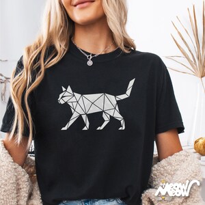 May include: Black t-shirt featuring a geometric cat design in white. The cat is composed of interconnected triangles and lines, creating a modern, minimalist aesthetic. The shirt has short sleeves and a crew neck.