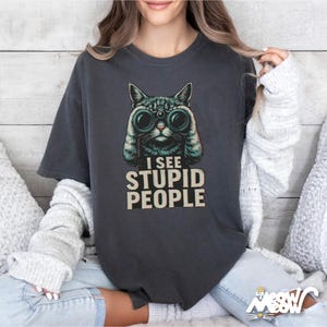 May include: Dark gray t-shirt featuring a cat with binoculars and the text "I SEE STUPID PEOPLE." The cat graphic is in shades of green and black. The shirt is a casual style.