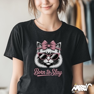 May include: Black t-shirt featuring a white cat face wearing pink sunglasses and a pink bow. The text "Born to Slay" is below the cat graphic in a pink script font. The shirt is a crew neck style.
