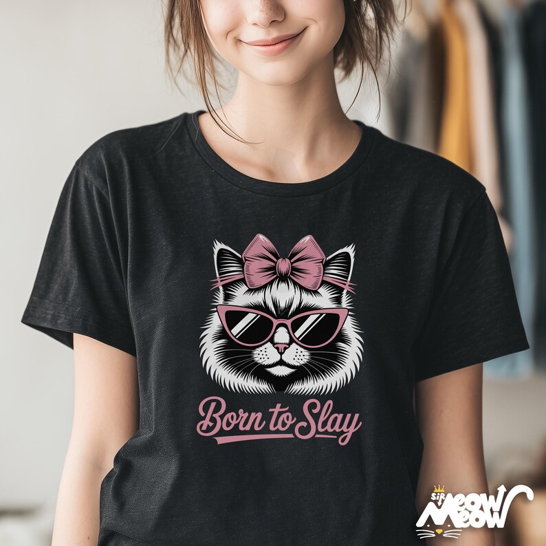 May include: Black t-shirt featuring a white cat face wearing pink sunglasses and a pink bow. The text "Born to Slay" is below the cat graphic in a pink script font. The shirt is a crew neck style.