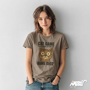 Puede incluir: Una camiseta marr&oacute;n con el texto "CAT GAME DON'T LOOK AT THE CAT GAME OVER" y una cara de gato de dibujos animados. La camiseta se lleva con unos vaqueros azules.