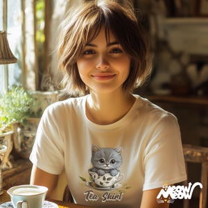 May include: A white t-shirt with a graphic of a gray kitten sitting in a teacup. The text "Tea Shirt" is printed below the image. The shirt is being worn by a person with short brown hair.