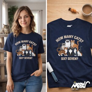 May include: Navy blue t-shirt with a graphic of several cats and the text "HOW MANY CATS? SIX? SEVEN?". The shirt features a crew neck and short sleeves. A woman is wearing the shirt in the image.