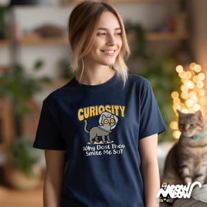 Op de afbeelding: Marineblauw t-shirt met een cartoon kat ontwerp en het woord "Curiosity". De zin "Why Dost Thou Smite Me So?" staat onder de kat. De afbeelding toont een kat met een helm. Een kat zit naast de persoon die het t-shirt draagt.