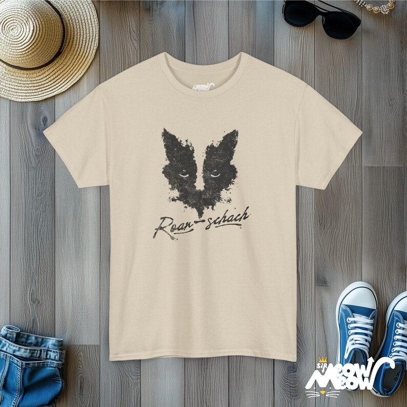 May include: A beige t-shirt with a black and white graphic of a badger's face and the text "Rorschach" in black.