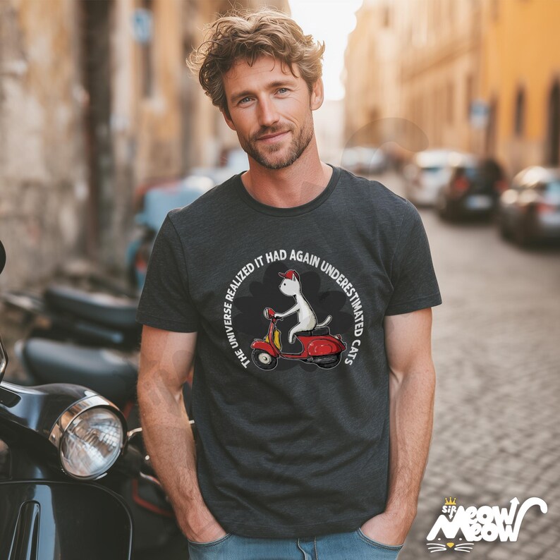 May include: Dark gray t-shirt featuring a cartoon cat riding a red scooter. The shirt has the text "THE UNIVERSE REALIZED IT HAD AGAIN UNDERESTIMATED CATS" in a circular design. The shirt also has the "Sir Meow Meow" logo.