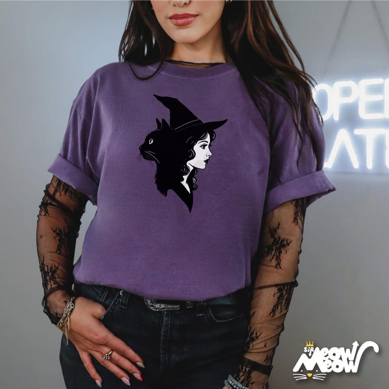 May include: A purple t-shirt featuring a black graphic of a witch's silhouette combined with a black cat. The shirt has rolled-up sleeves and is paired with black lace sleeves. The bottom right corner of the shirt has the text "Sip Meow Meow".
