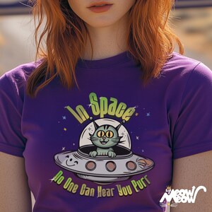 Może przedstawiać: Fioletowy T-shirt z zielonym kotem w statku kosmicznym. Tekst na koszulce brzmi "In Space No One Can Hear You Purr".