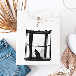 May include: White Comfort Colors sweatshirt featuring a black illustration of a cat sitting in a bay window. The window design is detailed, with a cityscape visible through the glass. A silver necklace and a pair of jeans are also visible.