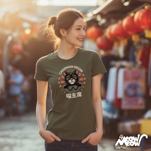 May include: Olive green t-shirt featuring a graphic of a black cat wearing a military-style uniform and a red star. The text "CHAIRMAN MEOW" and Chinese characters are above and below the cat illustration. The shirt has short sleeves.