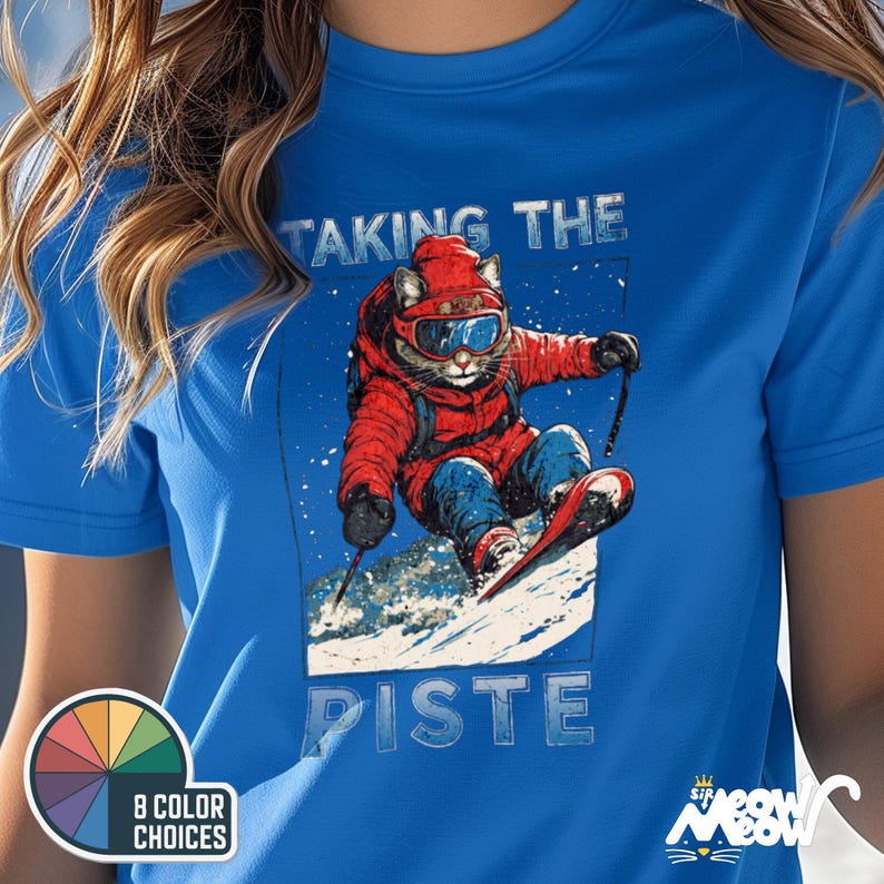 K&ouml;nnte beinhalten: Ein blaues T-Shirt mit einem Aufdruck eines katzenartigen Wesens, das Ski f&auml;hrt. Der Text "Taking the Piste" ist auf dem Shirt aufgedruckt.