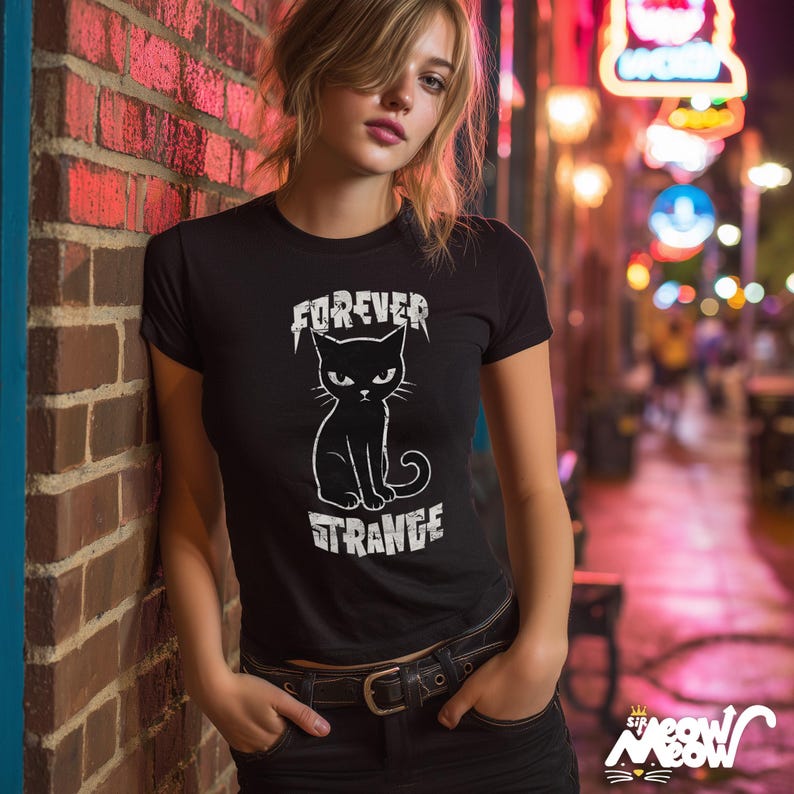 May include: Black t-shirt with the words "FOREVER STRANGE" and a graphic of a black cat with an angry expression. The shirt is short-sleeved and has a crew neck. The model is wearing black jeans and a belt.