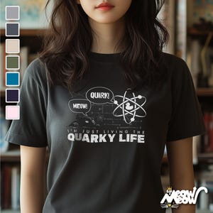 May include: A dark gray t-shirt with a white graphic of a cartoon duck in an atom. The text on the shirt reads "I'm just living the quirky life" with the words "quark!" and "meow!" in speech bubbles.