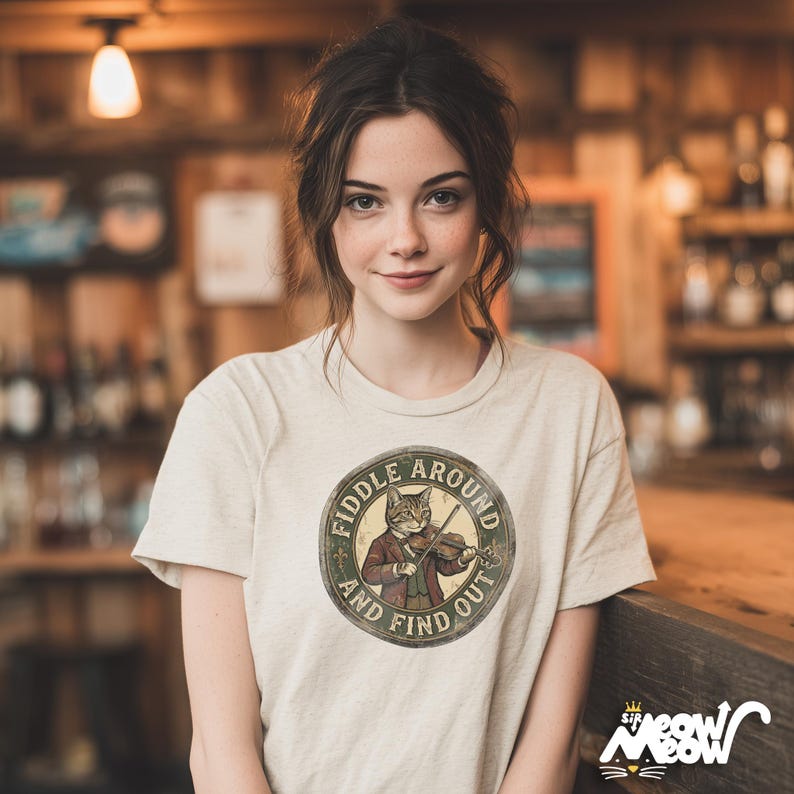 May include: A light beige t-shirt featuring a circular graphic with a cat playing a violin, the text "FIDDLE AROUND AND FIND OUT". The shirt has short sleeves and a crew neck. The brand logo "Sit Meow Meow" is at the bottom right.
