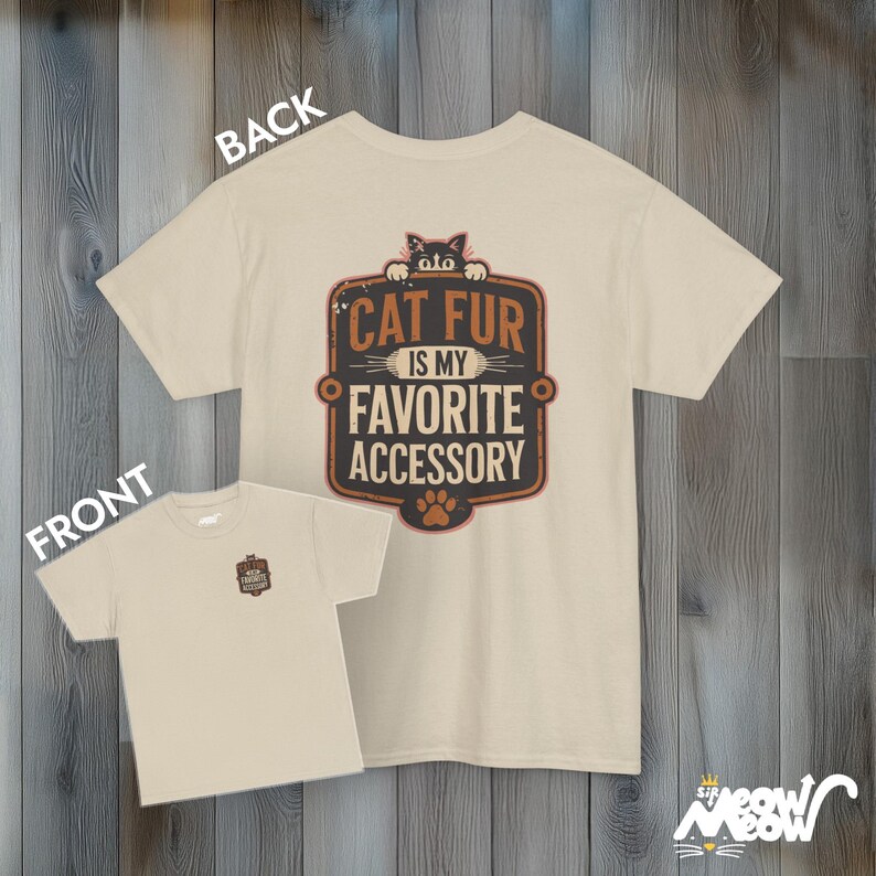 May include: A beige t-shirt with a vintage-style graphic that says "Cat fur is my favorite accessory". A black cat peeks over the top of the graphic. The t-shirt is shown on a wooden background.
