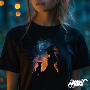 May include: A black t-shirt featuring a colourful graphic of a witch and a cat standing in front of a crescent moon. The graphic is surrounded by a watercolour-like background.