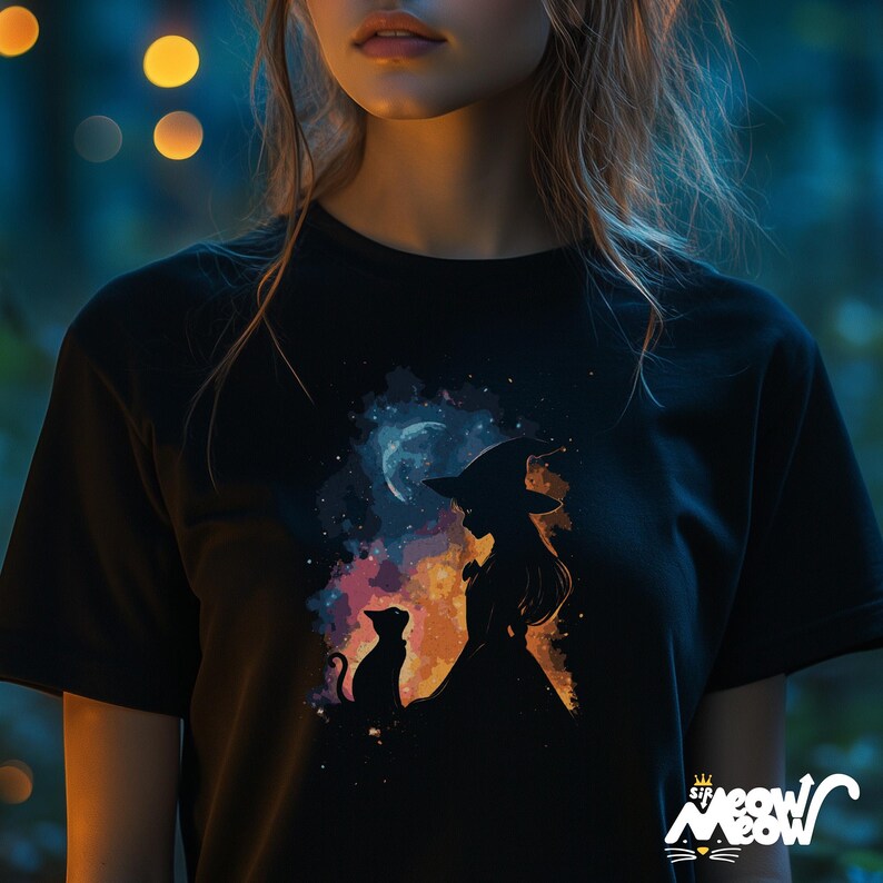 May include: A black t-shirt featuring a colourful graphic of a witch and a cat standing in front of a crescent moon. The graphic is surrounded by a watercolour-like background.