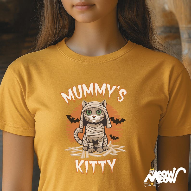 Pode incluir: Camiseta amarelo mostarda com um gr&aacute;fico Mummy Kitty. O design apresenta um gato de desenho animado enrolado em bandagens, com olhos verdes e as palavras "Mummy's Kitty". Morcegos pretos e um pequeno logotipo tamb&eacute;m est&atilde;o presentes.